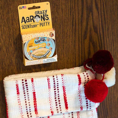 Package of Gingersnapper SCENTsory™ Thinking Putty® next to a stocking on a wood table.